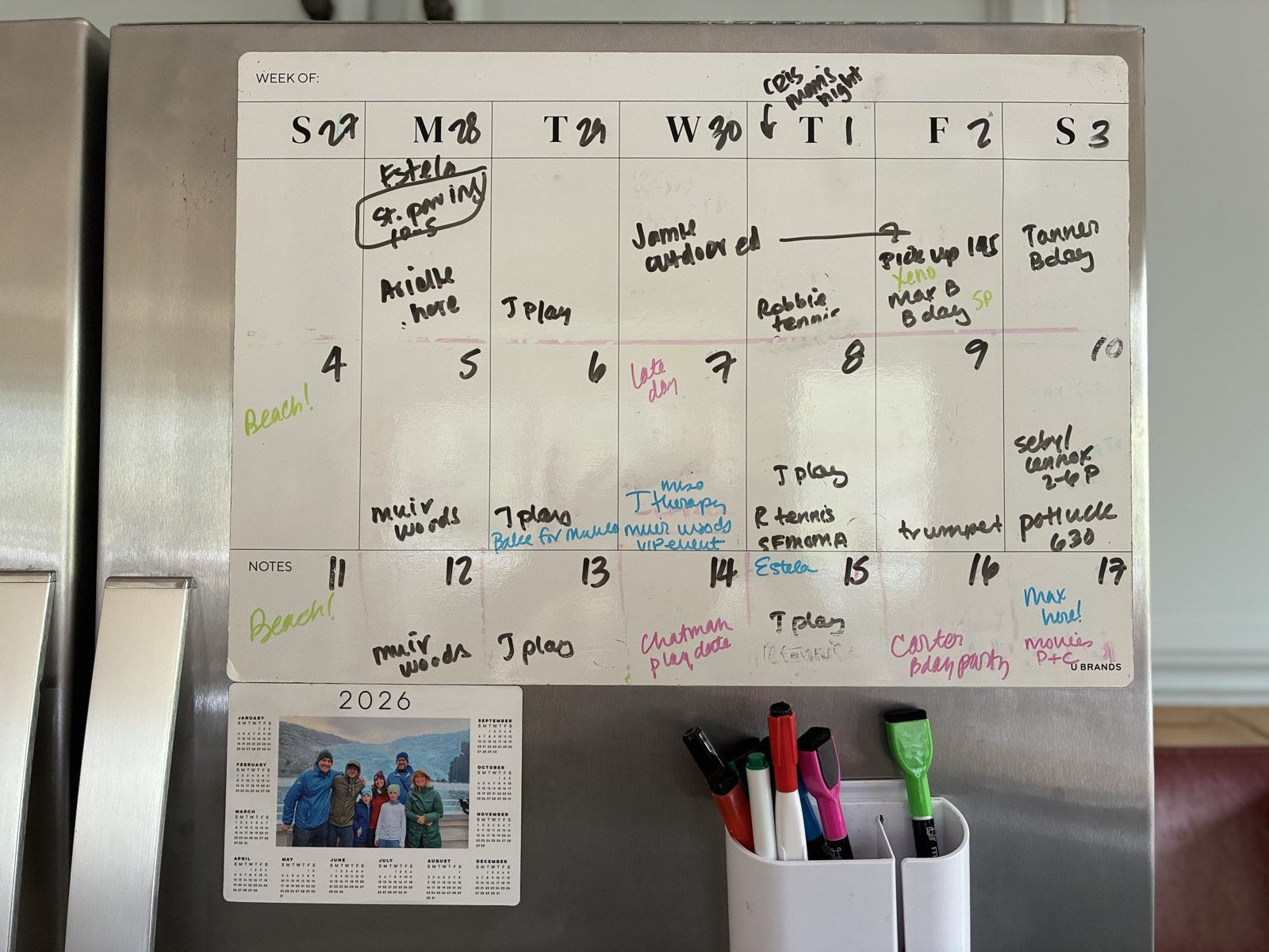 Our real fridge: a whiteboard week calendar full of handwritten plans in colored marker, a cup of dry-erase markers, and a small family photo calendar